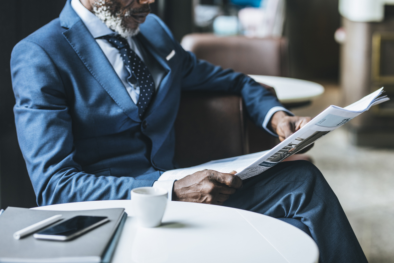 African businessman wearing suit and tie sitting at coffee shop and reading daily newspaper.