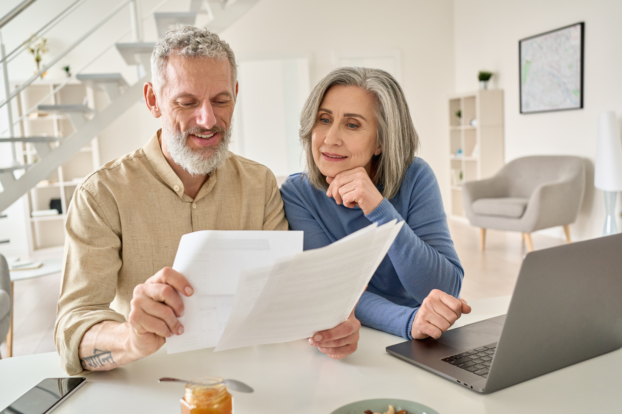 Middle aged senior old couple holding documents reading paper bills paying bank loan online, calculating pension fees, payments, taxes, planning family retirement money finances using laptop at home.