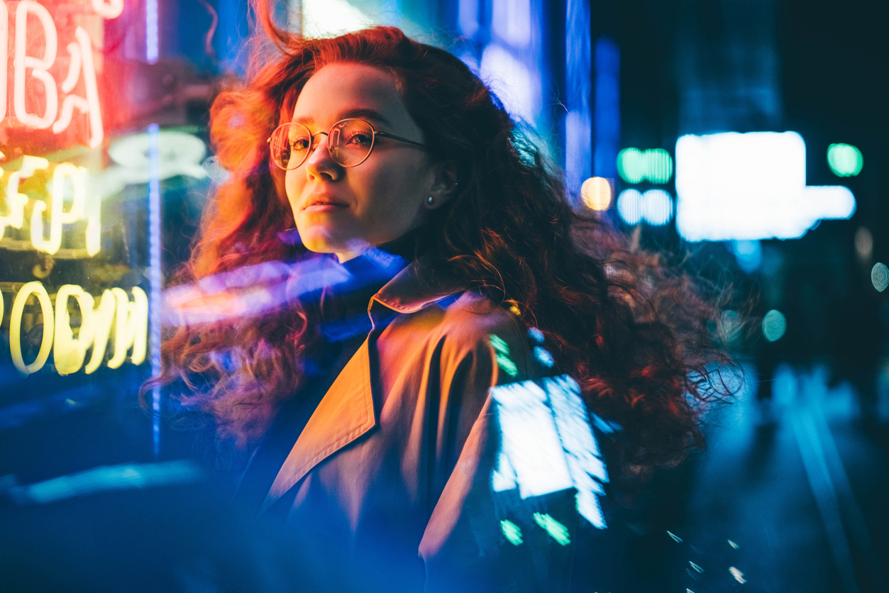 Curly haired young woman tourist with light makeup in glasses looks around standing near bar with colorful neon sign against night megalopolis