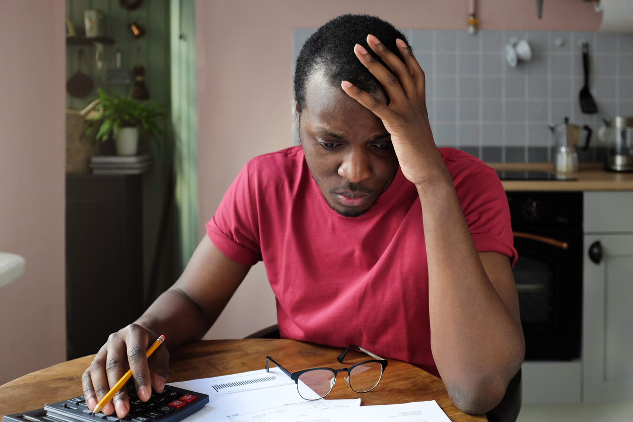 young anxious worrying african american man looks desperately to bills, faces problems with paying taxes, holding head with one hand