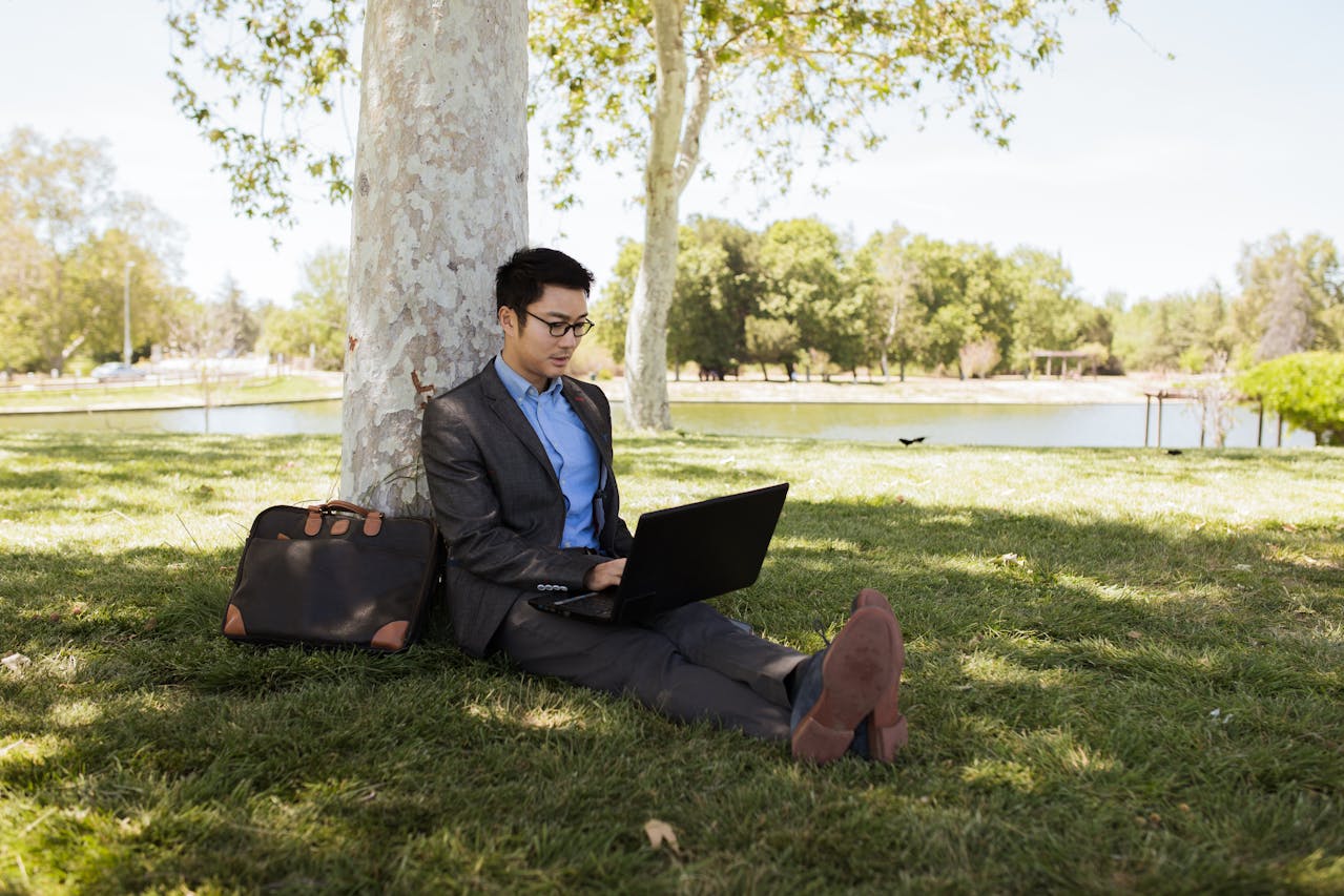 Young man on a laptop sat in a park leaning against a tree.