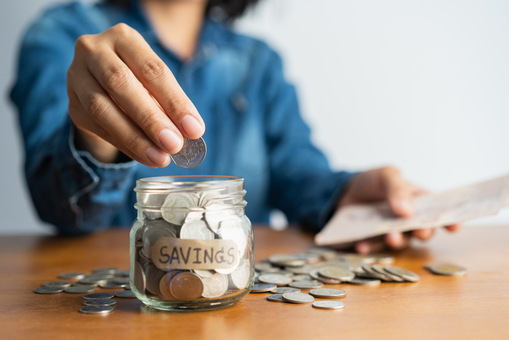 the woman hand is putting a coin in a glass bottle and a pile of coins on a brown wooden table,investment business, retirement, finance and saving money for future concept.