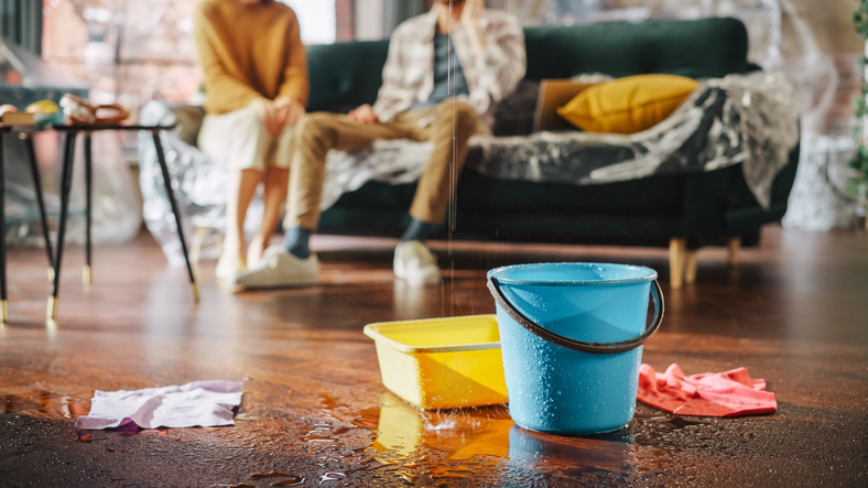 roof is leaking, pipe rupture at home: water drips into buckets in living room. angry couple in background calling insurance company, screaming into phone in frustration, trying to find plumber