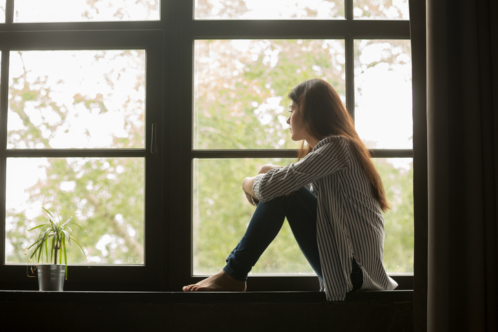 thoughtful girl sitting on sill embracing knees looking at window