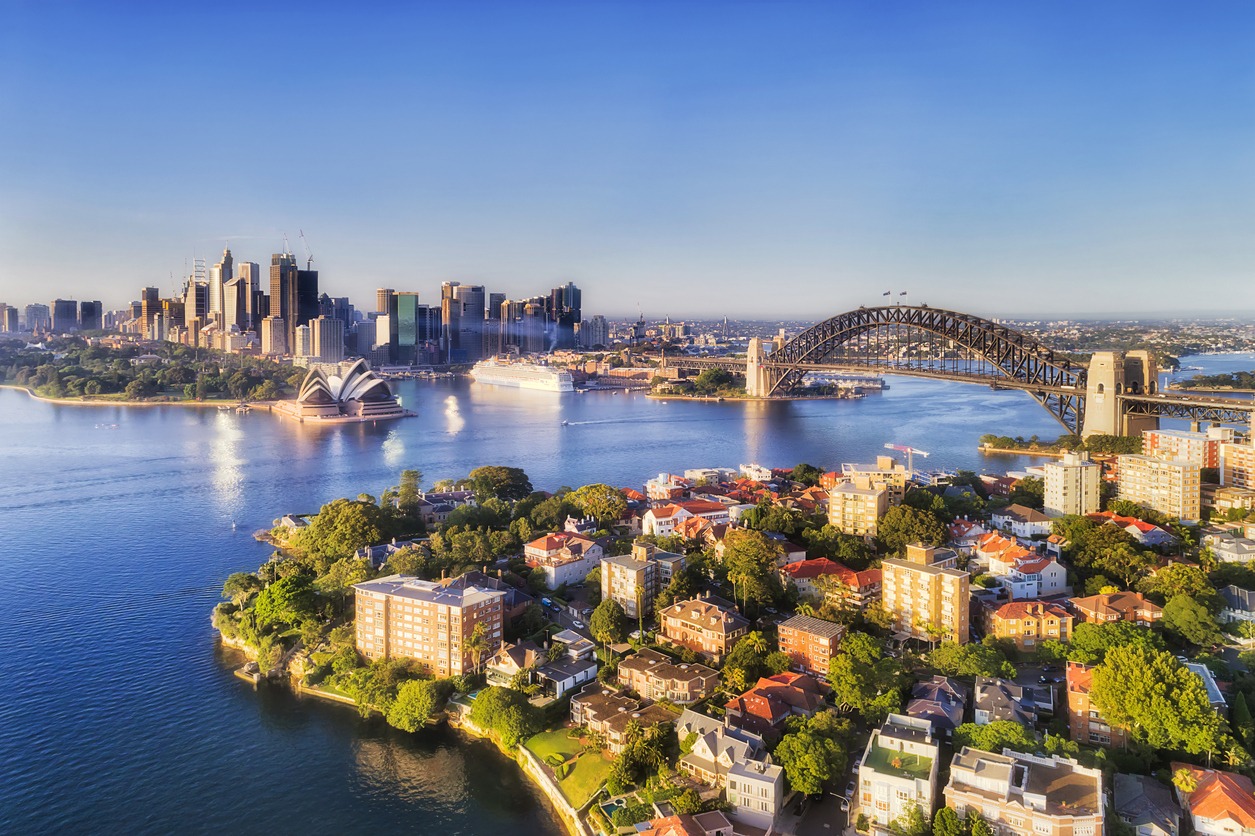 Still blue waters of Sydney harbour between city CBD landmarking builidings connected by the Sydney Harbour bridge to North Shore wealthy suburbs with Kirribilli in forefront of aerial view.