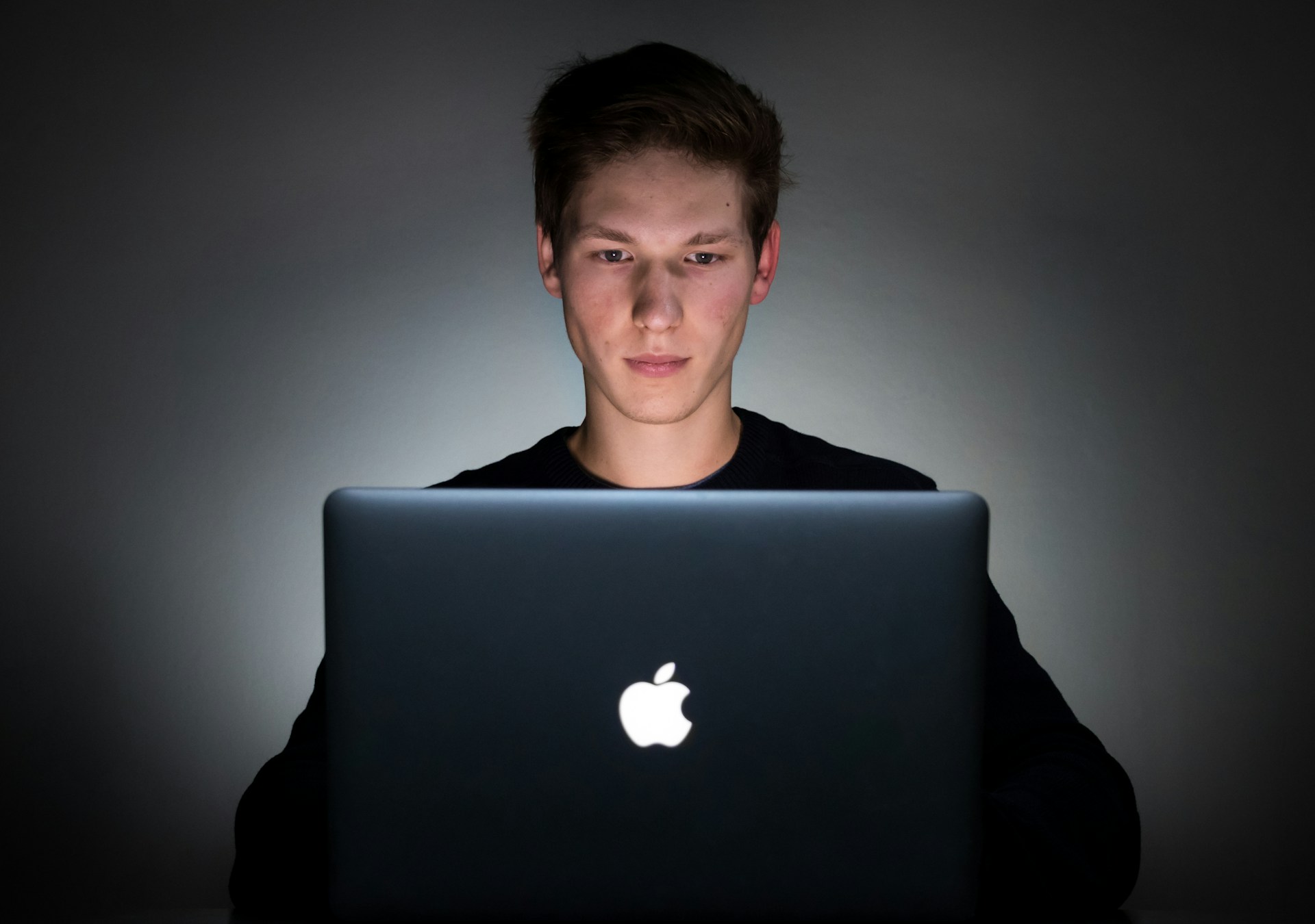 Young man backlit by light in a dark room working on a mac laptop.