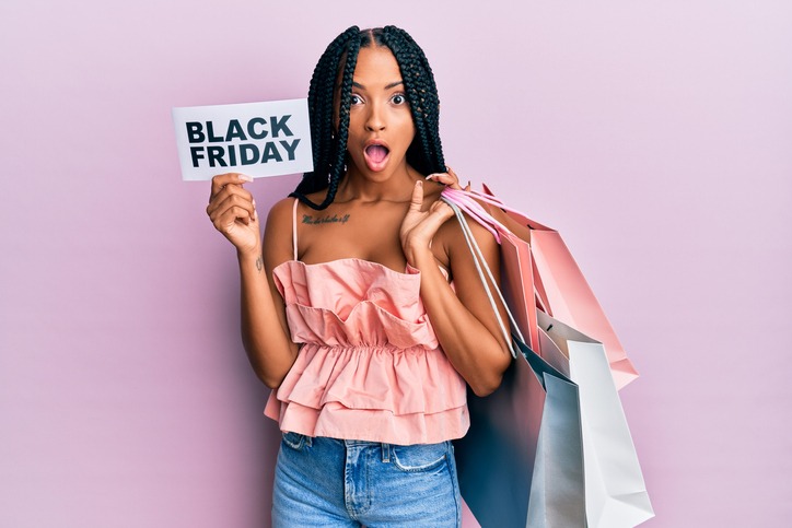 beautiful hispanic woman holding shopping bags and black friday banner afraid and shocked with surprise and amazed expression, fear and excited face.