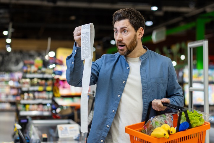 shocked caucasian man with rounded eyes and open mouth looking at extremely long bill while holding orange food basket. amazed buyer doing grocery shopping and getting surprised by prices