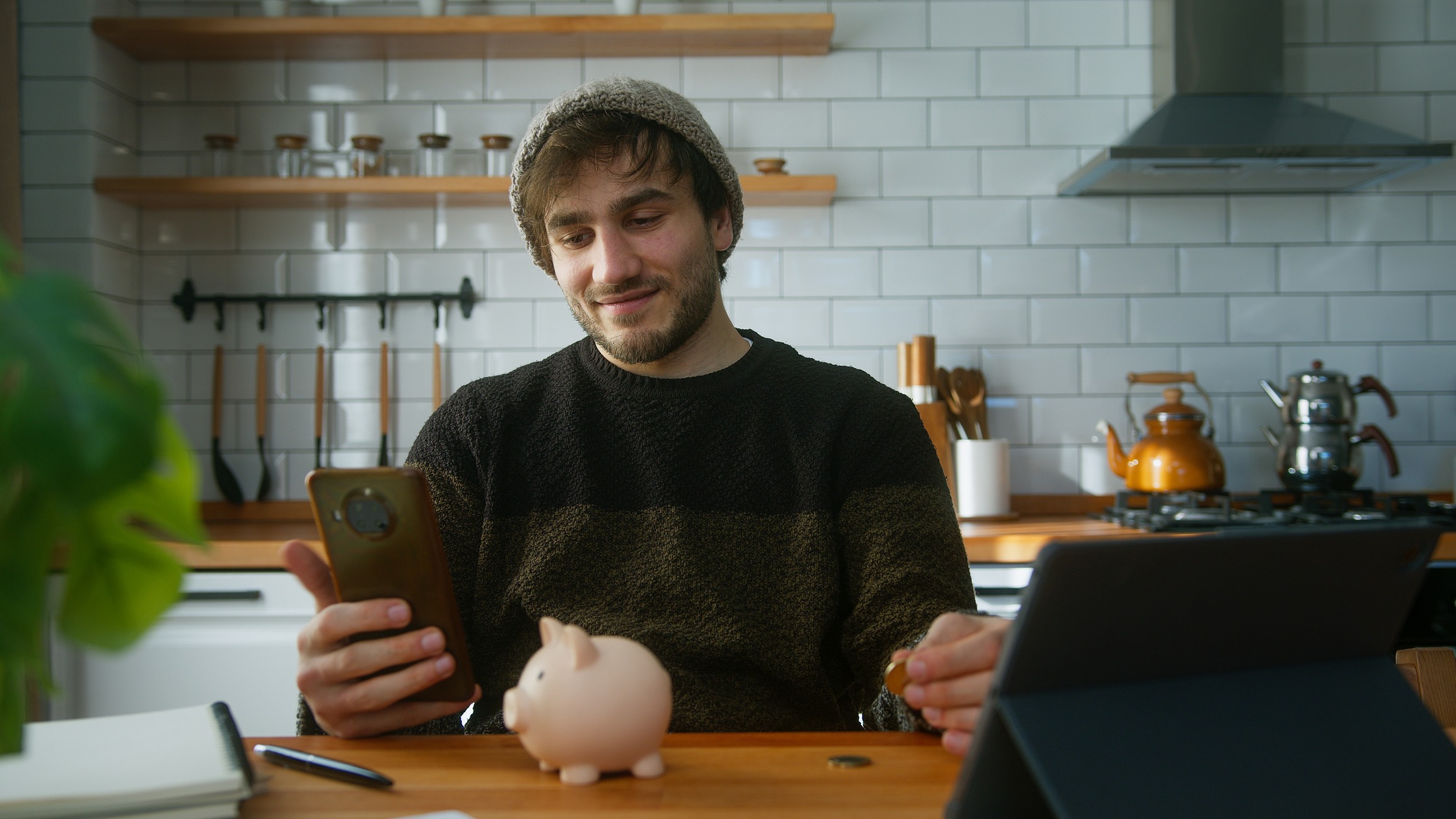 happy man with beanie sitting in modern kitchen at home while holding smartphone in his hand looking at the camera and puts couple of coin into the piggy bank